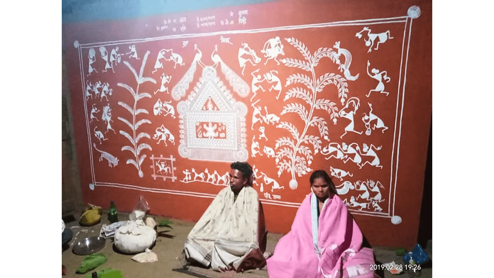 A traditional Warli painting done in white over a red ochre background. Panchasiriya is in the dev chauk in the bottom left of the picture. there is a lagna chauk in the centre.
